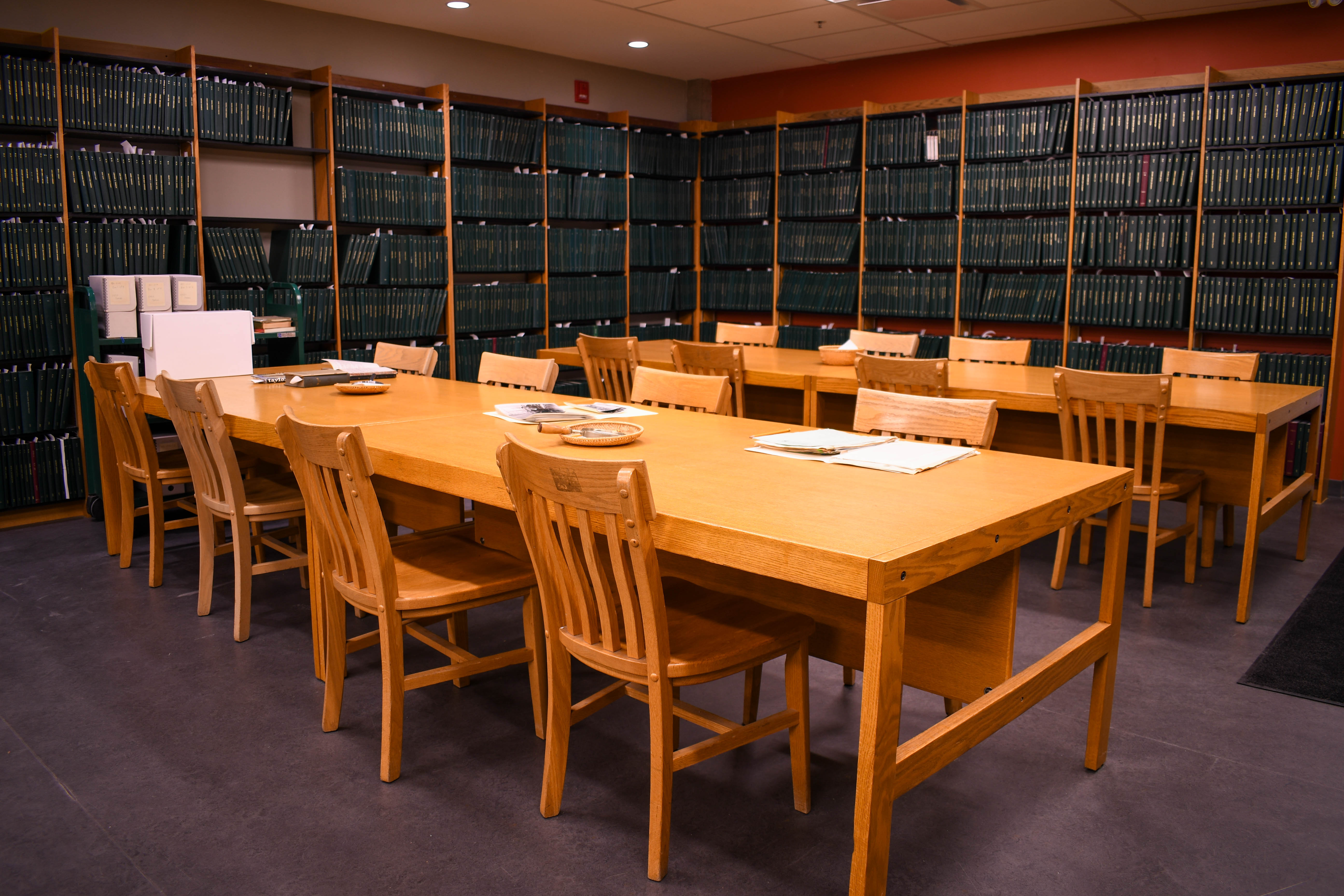 Tables and chairs in an archival reading room. There are boxes and other archival material on the tables and green books lining the walls.
